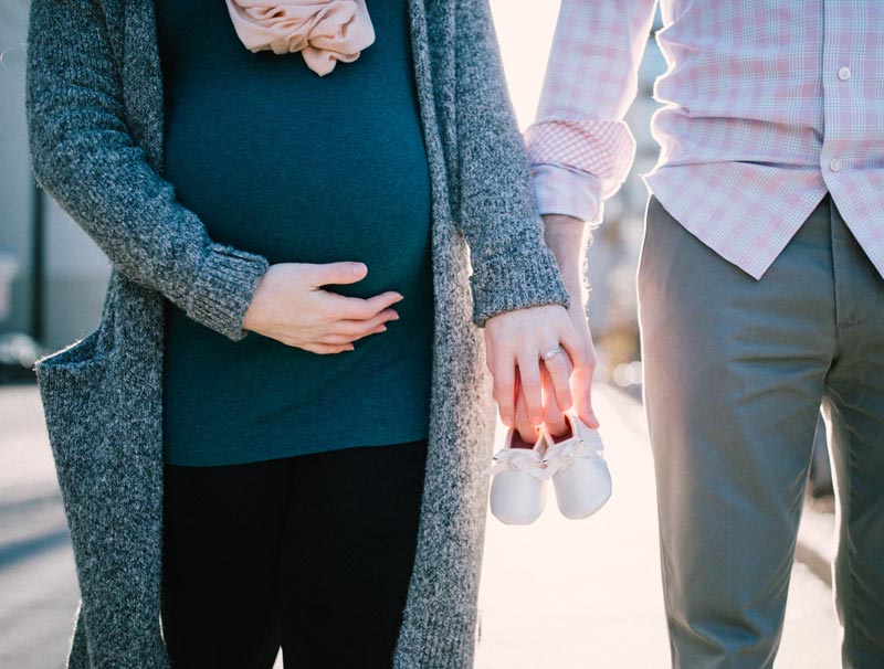 A Pregnant Lady Walking With a Man Holding Baby Shoes